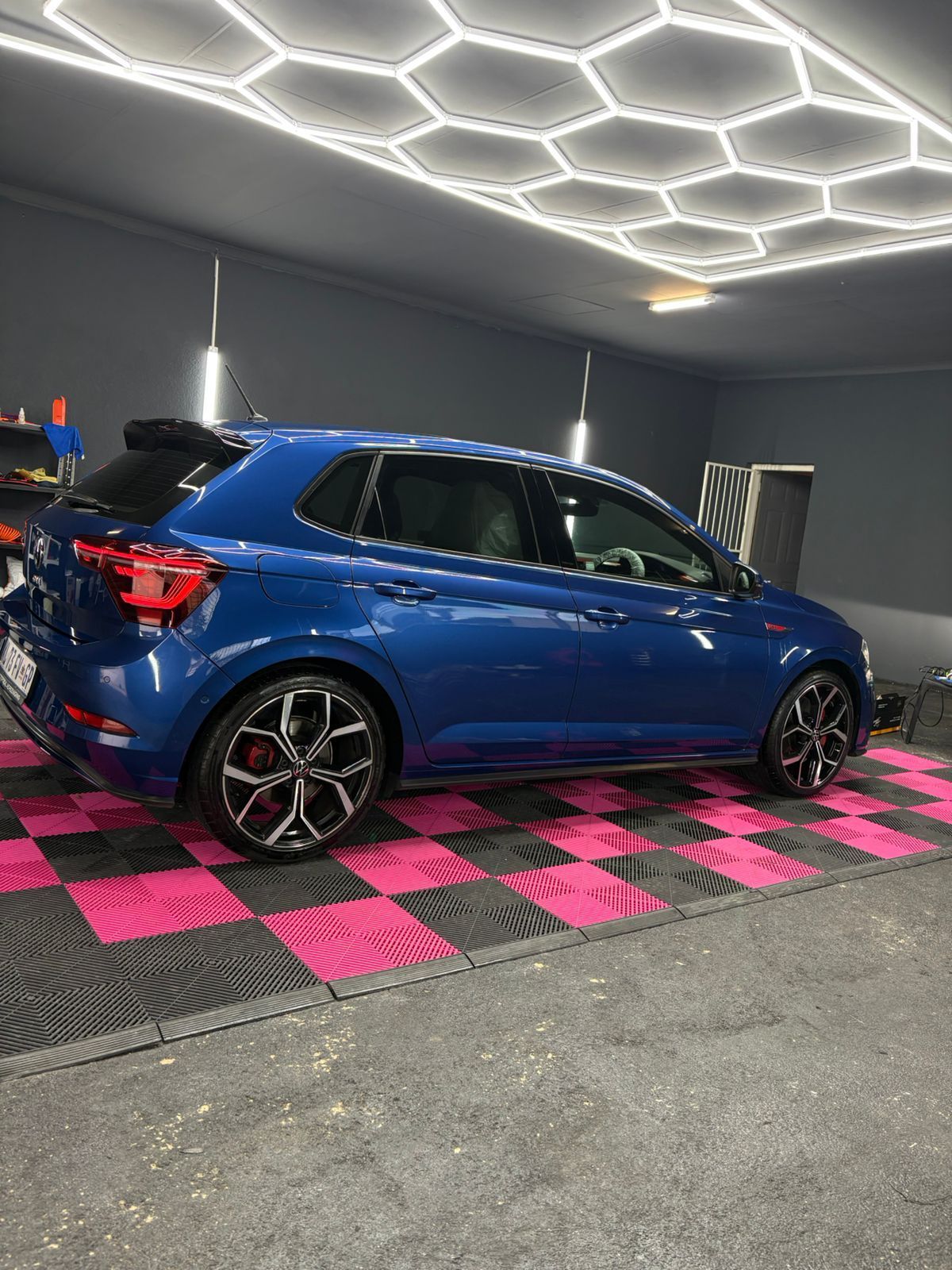 Blue car in a garage with black and pink checkered floor and hexagonal ceiling lights.