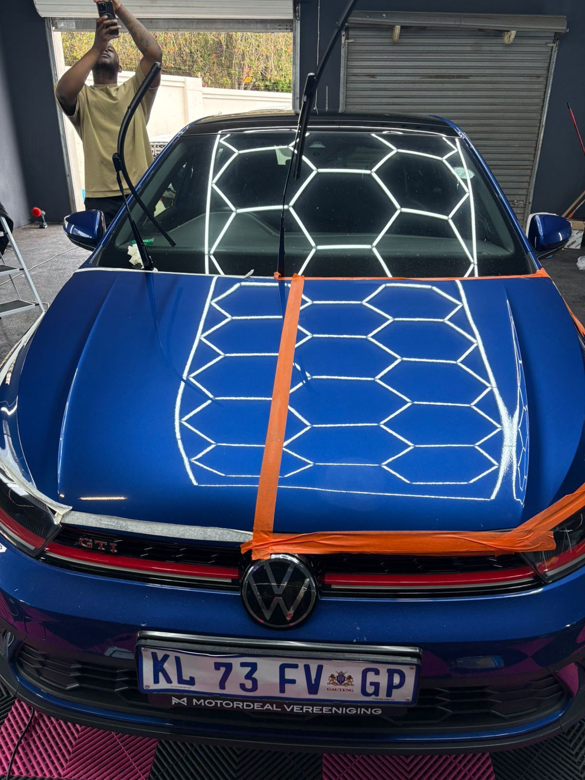 Blue car with orange tape down the center of the hood, a person applying something to the car.