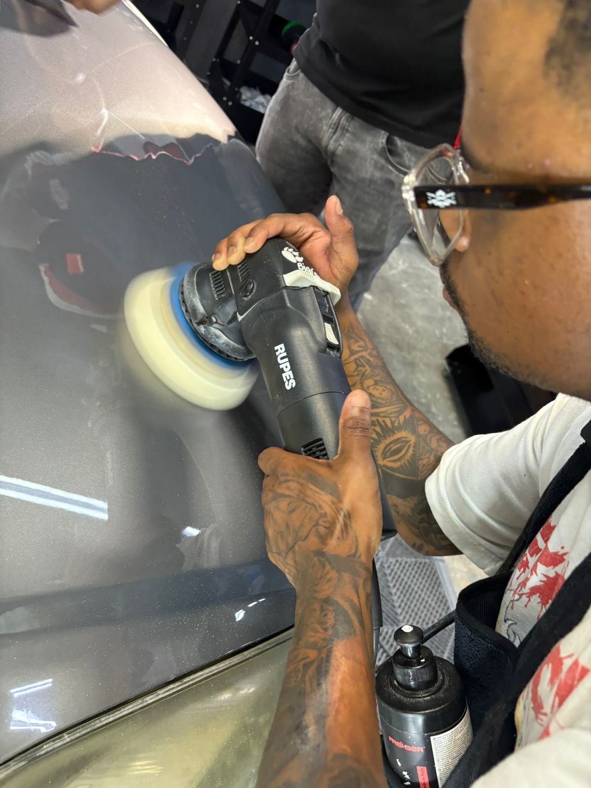 Man polishing a car with an electric buffer in a well-lit workshop.