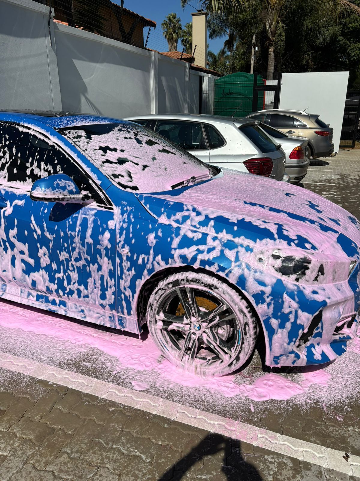 Blue car covered in pink foam, possibly being washed outdoors.