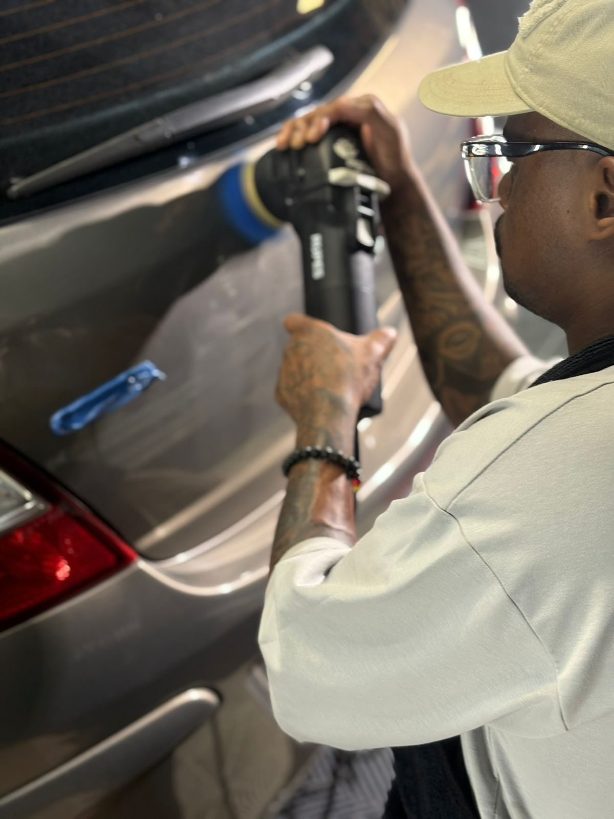 Person buffing car with a machine; wearing a white cap, glasses, and a light-colored shirt.