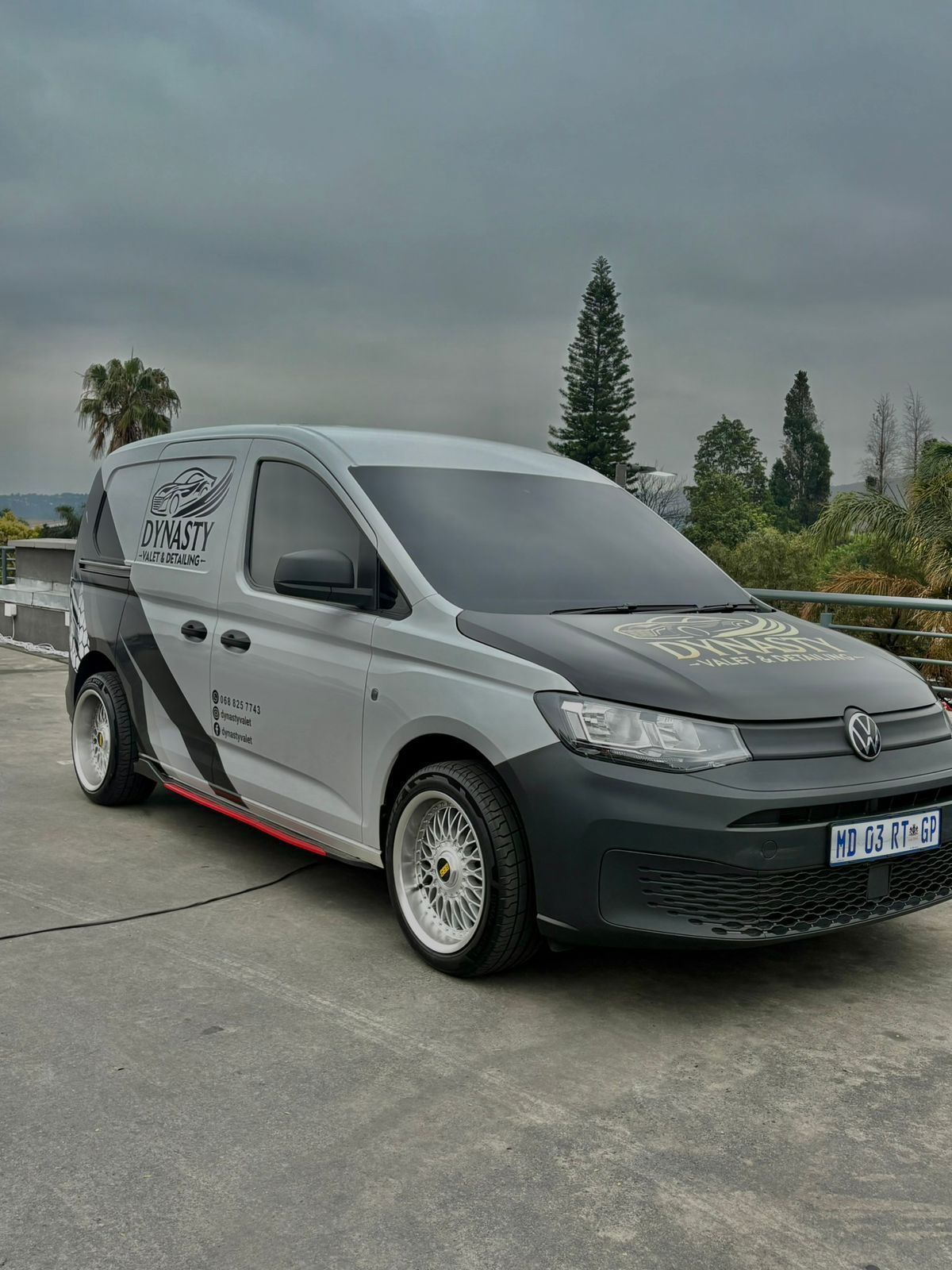 A modified gray and black Volkswagen van with custom wheels, parked outdoors on a cloudy day.