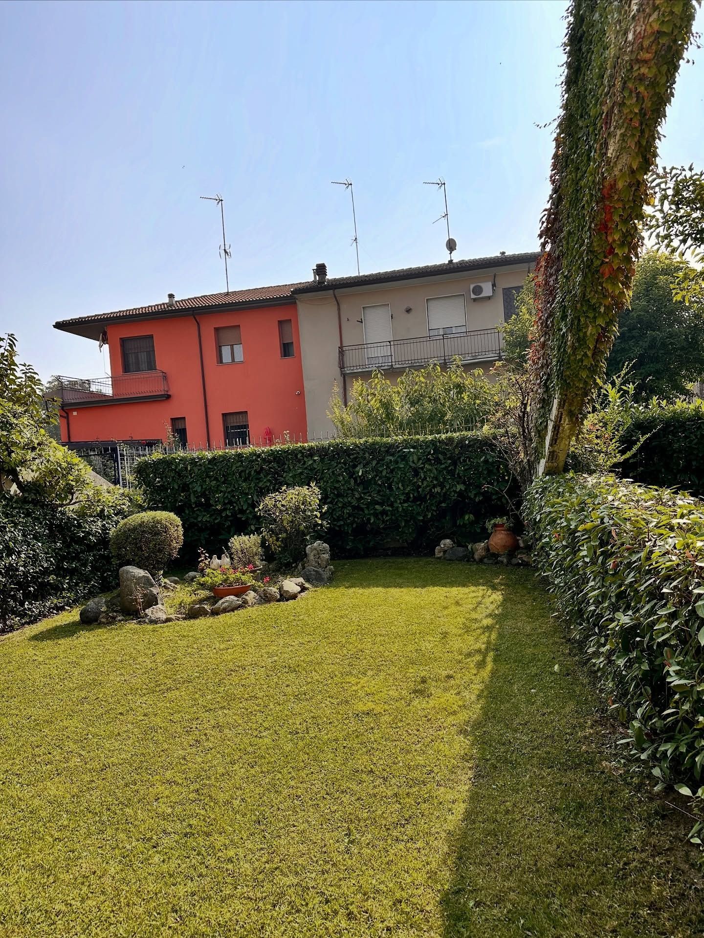 Un prato verde brillante, incorniciato da un'alta siepe, conduce a una casa a due piani con una facciata rossa e una sezione grigia e deteriorata dagli agenti atmosferici.