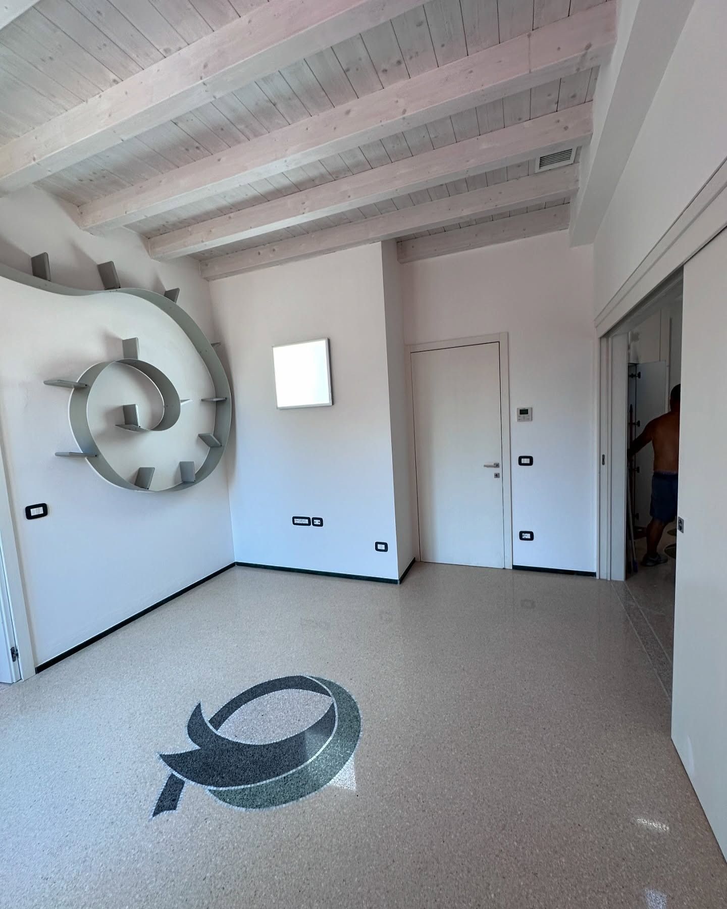 Una stanza con pareti bianche, una mensola a spirale, pavimento in terrazzo con un logo e una porta che mostra una figura parziale.