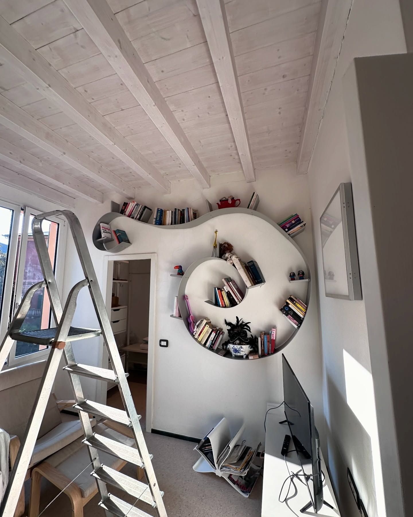 Una libreria a spirale installata a parete in una stanza con soffitto a travi bianche, con una scala a pioli in primo piano.