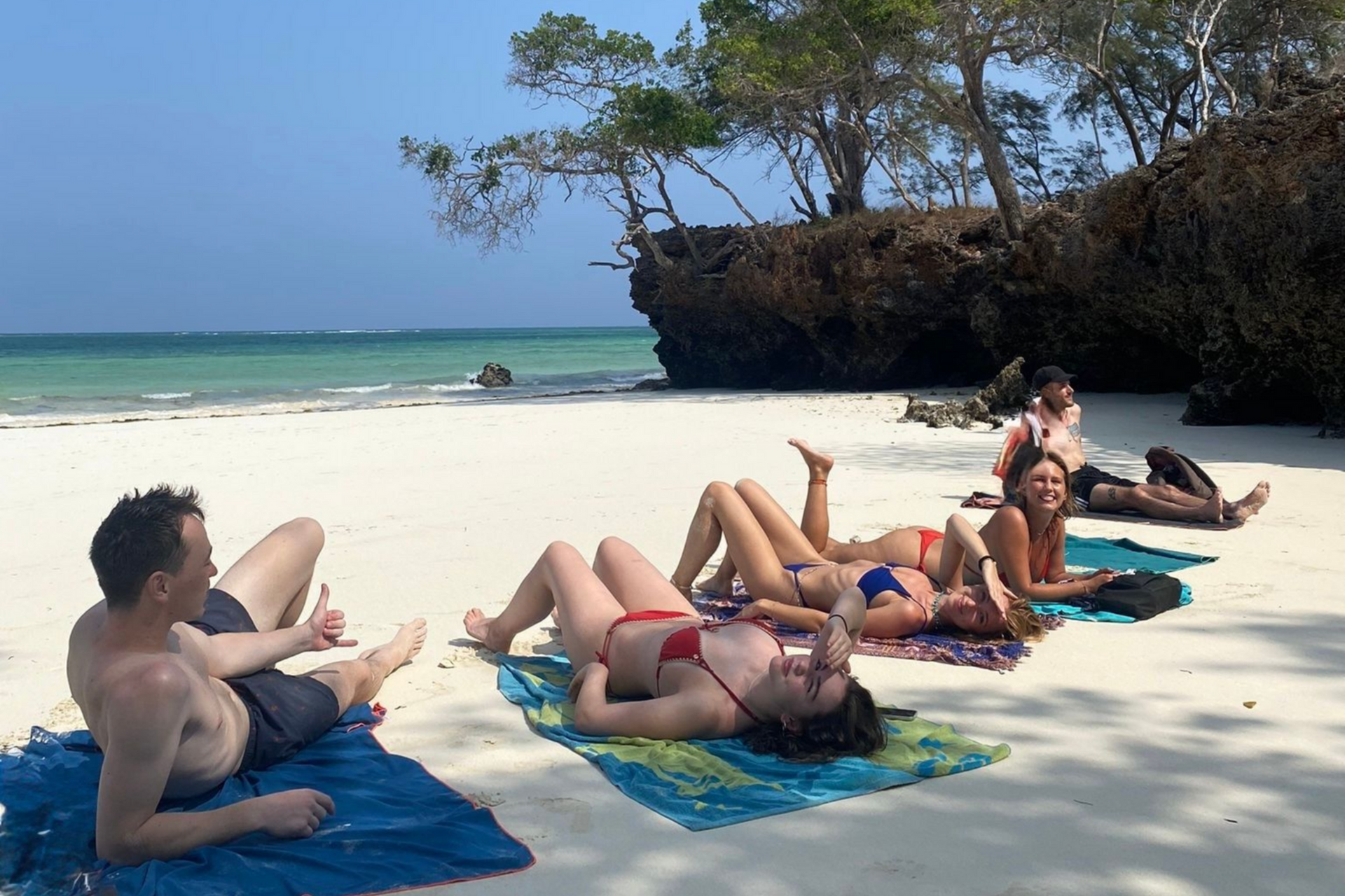 Woman sitting on white sand beach, facing ocean, wearing yellow top and denim shorts.