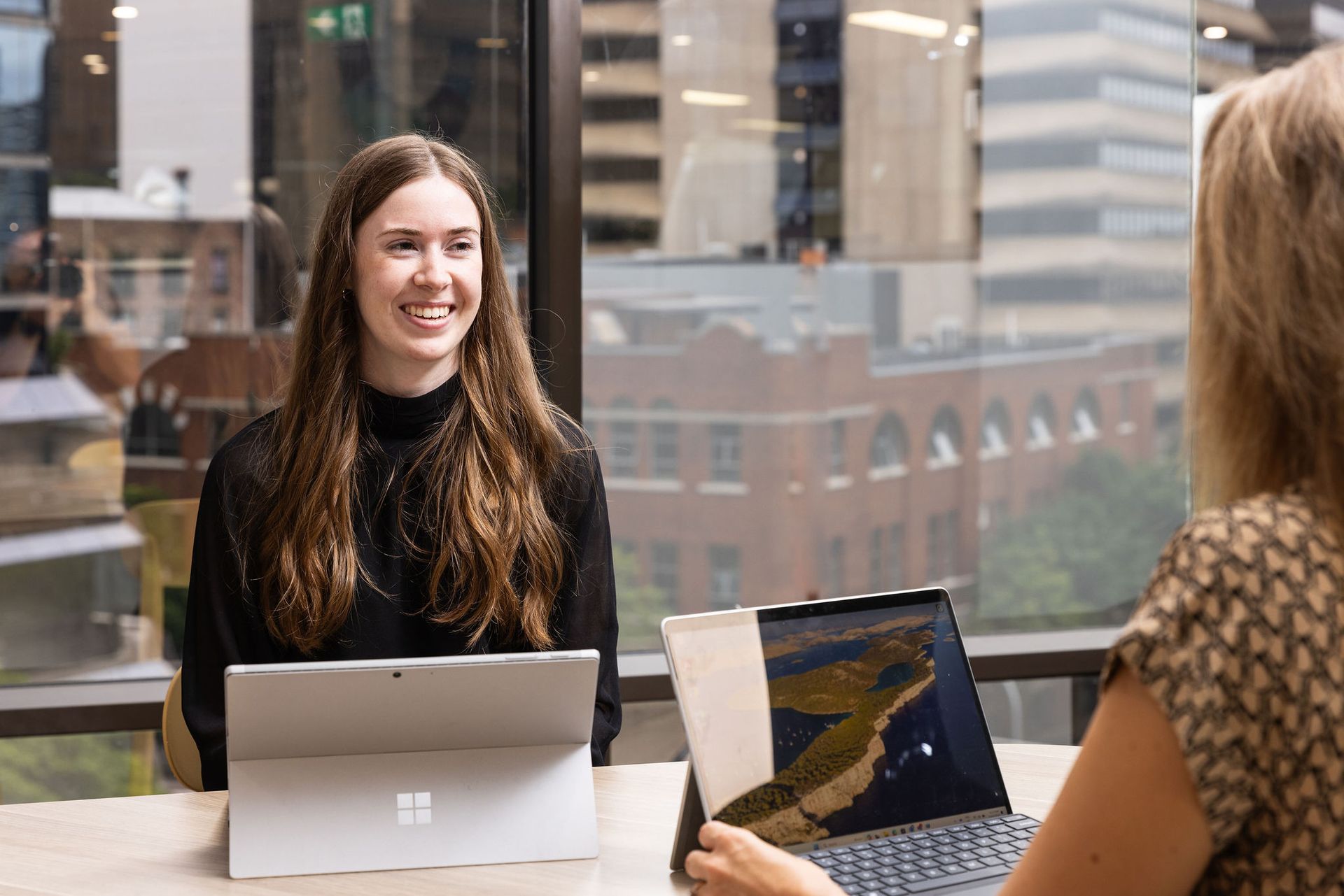 Two women are sitting at a table with laptops and talking to each other.