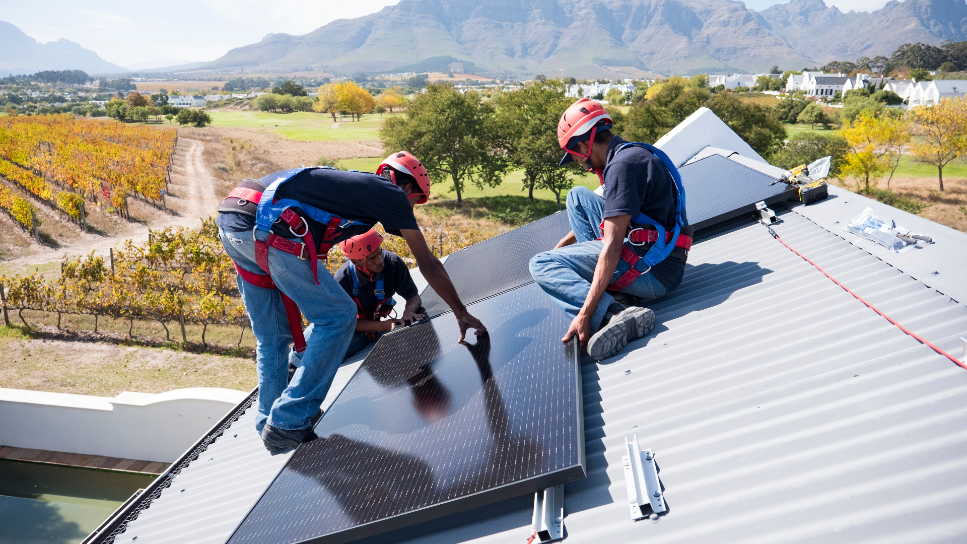 Workers in hard hats installing solar panels on a roof, mountains in the background.