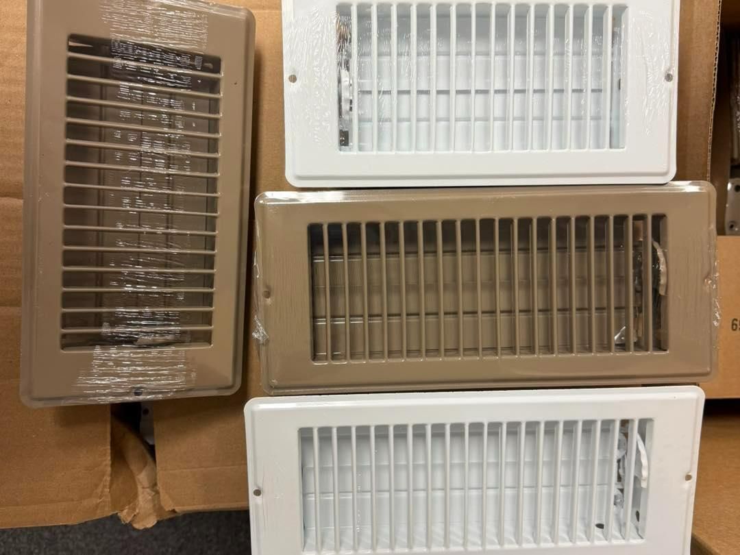 Four metal air vents in various colors, including beige and white, stacked on cardboard boxes.