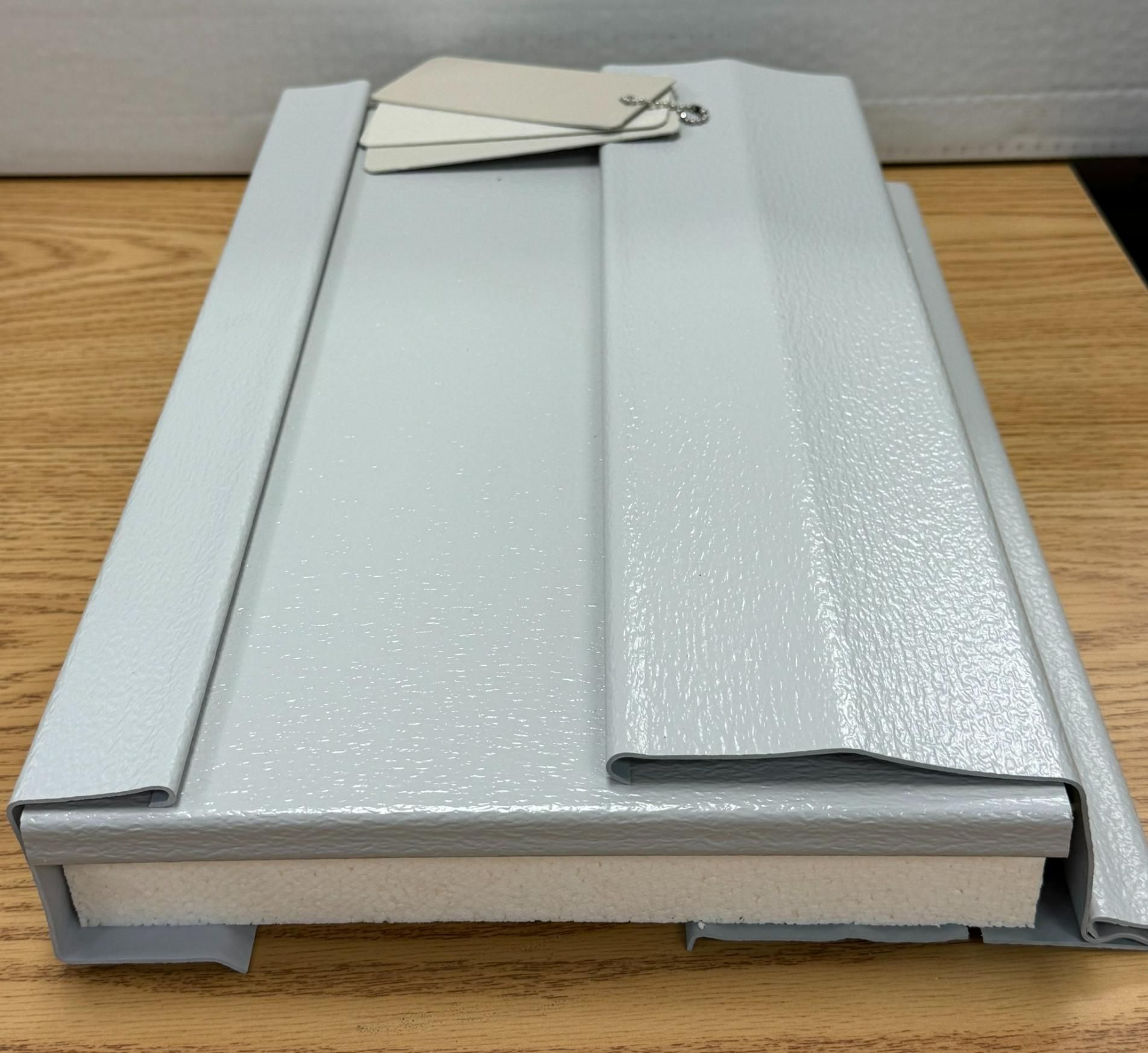 A light gray metal sample on a wooden table with paint color swatches attached.