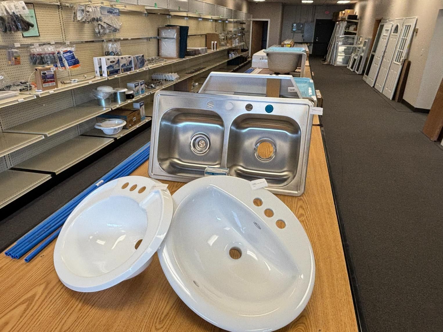 Two white bathroom sinks and a stainless steel kitchen sink on a wooden table in a store.