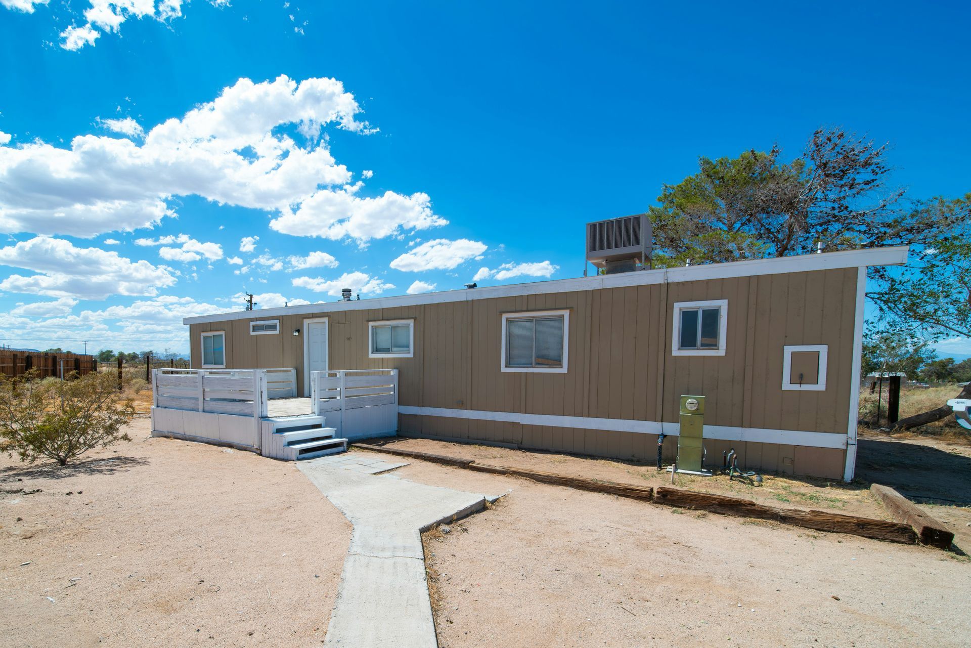 Tan mobile home in desert landscape under a blue sky with fluffy clouds.