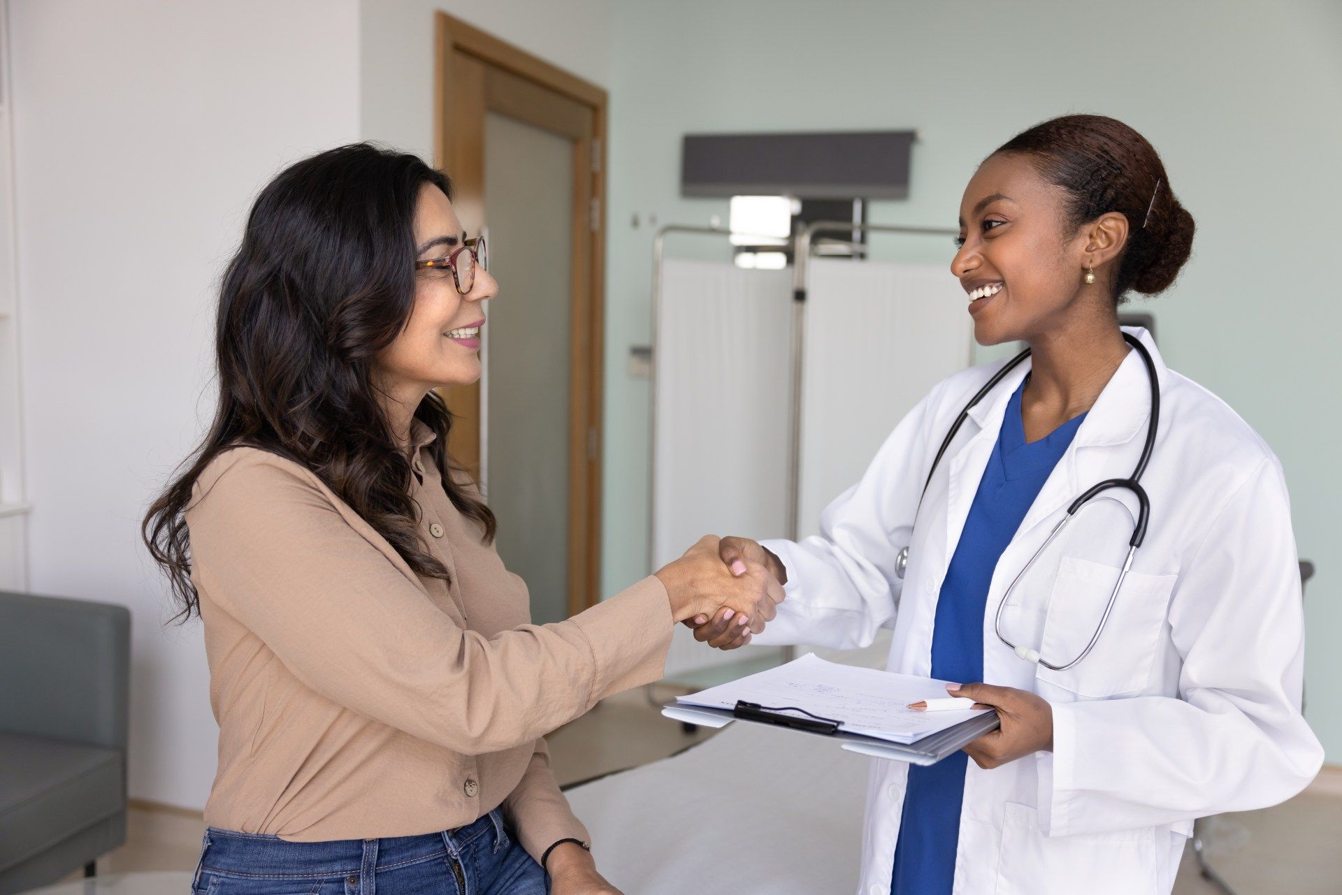 Patient shaking hands with a doctor in a clinic exam room, smiling and holding a clipboard.