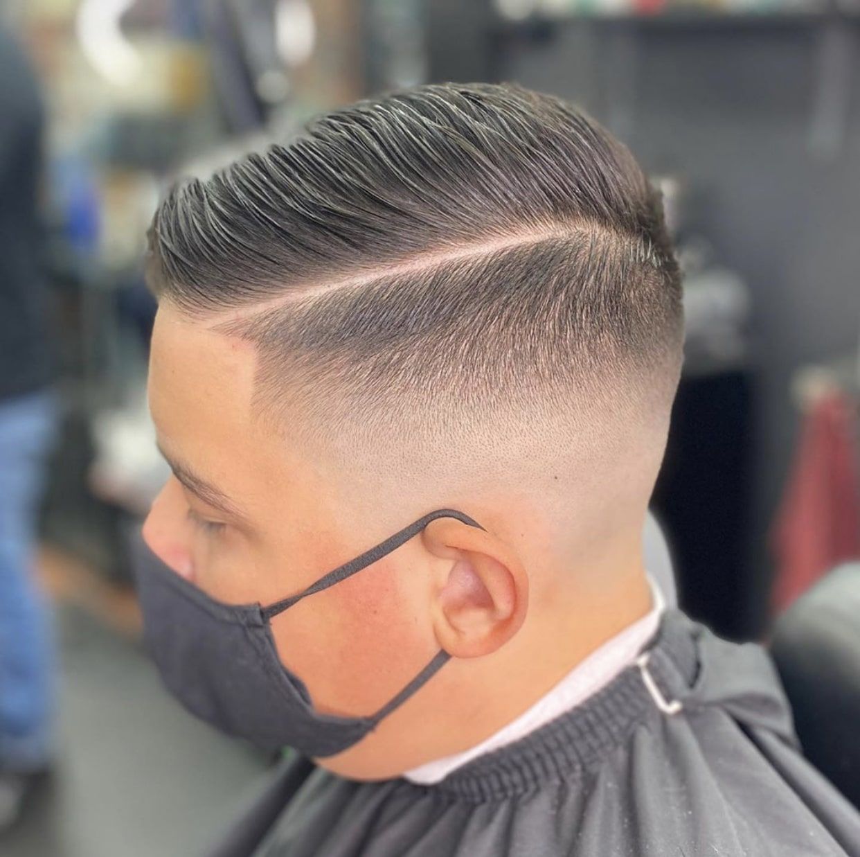 A man wearing a mask is getting his hair cut at a barber shop showing the low-fade comb over style.