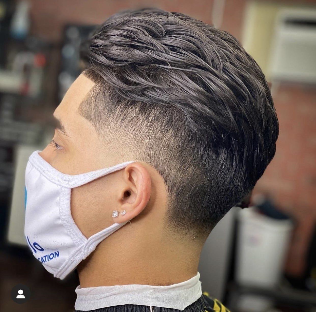 A man wearing a face mask is getting his hair cut by a barber showing his low-taper slick back style.