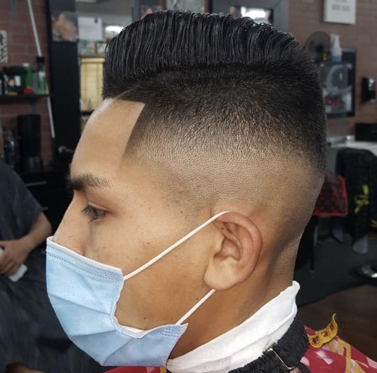 A man wearing a face mask is getting his hair cut with a mid-fade comb over style.