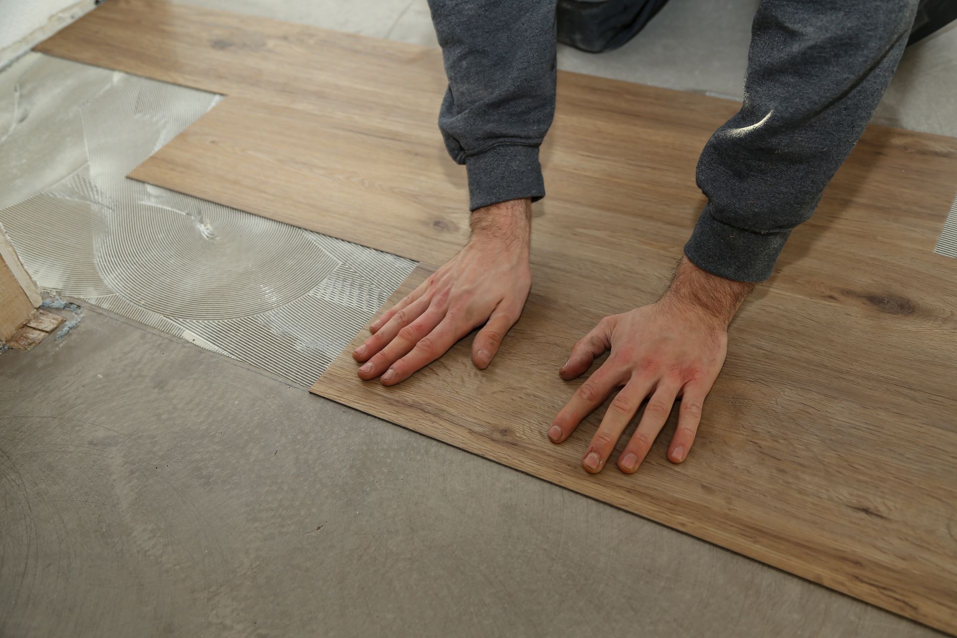 Person installing wood-look flooring, hands pressing a plank into adhesive on the subfloor.