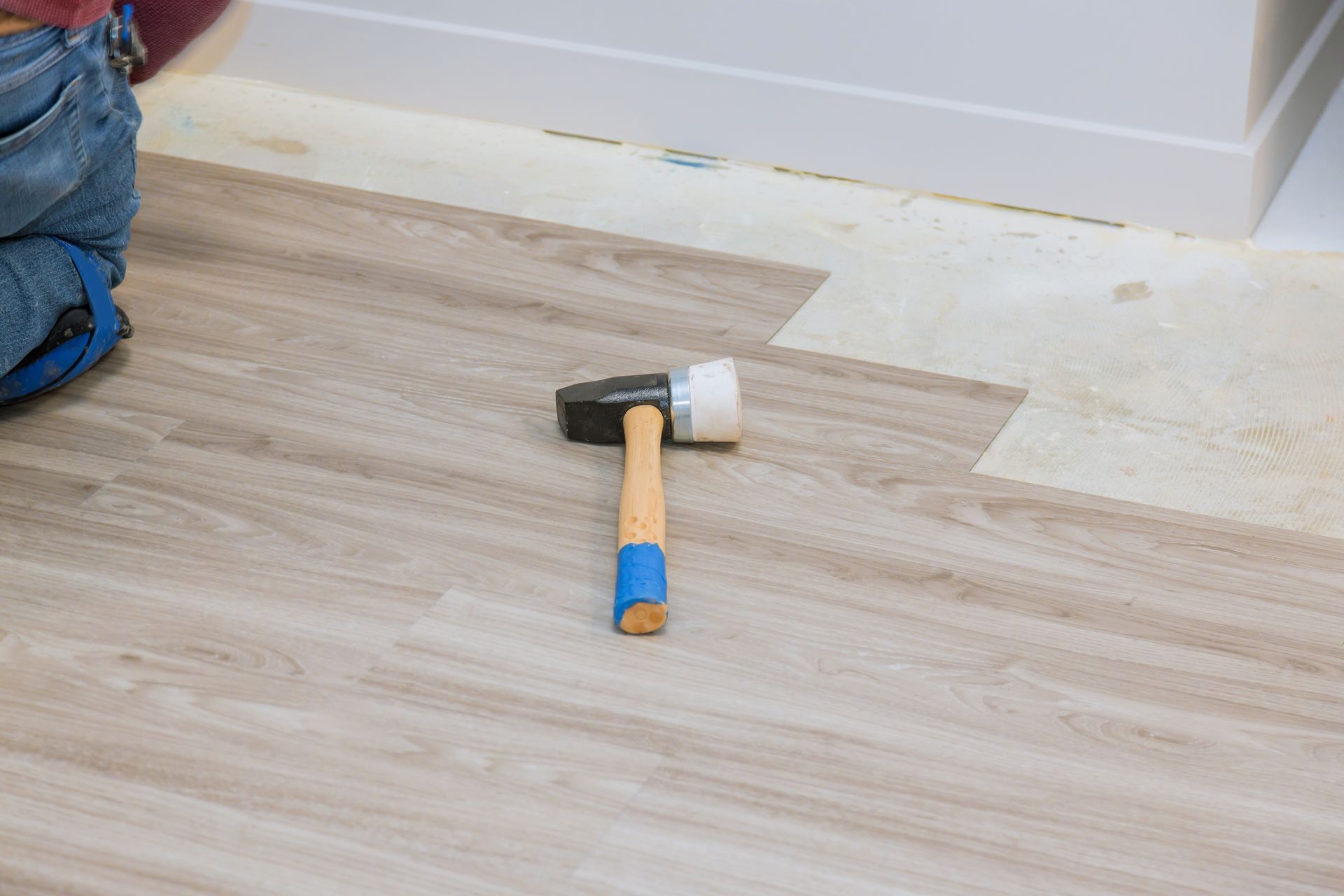 Hammer on wooden floor, near installed flooring and baseboard. Person installing flooring in the background.