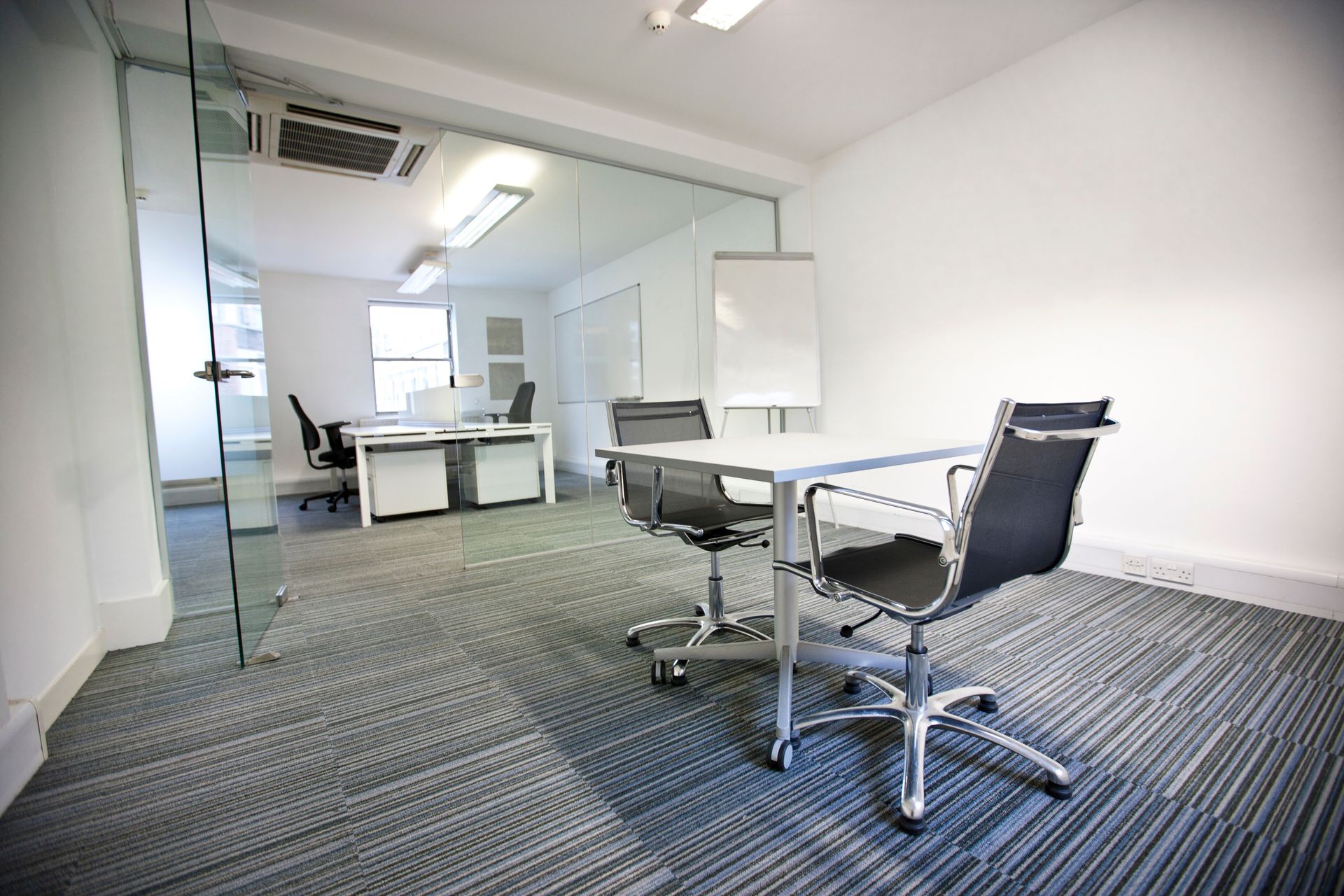 Modern office space with a meeting table and chairs, visible through a glass door. Grey carpet and white walls.
