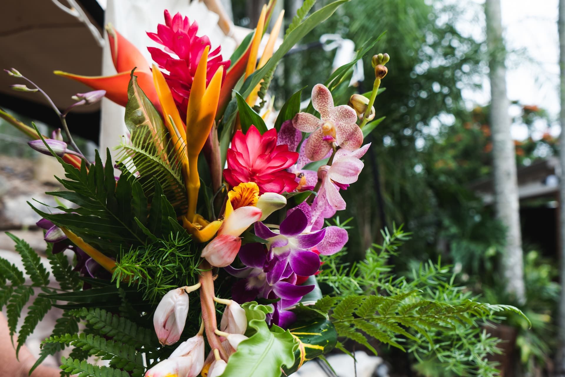 Colorful tropical flowers in a bouquet, including orchids, ginger flowers, and fern leaves.