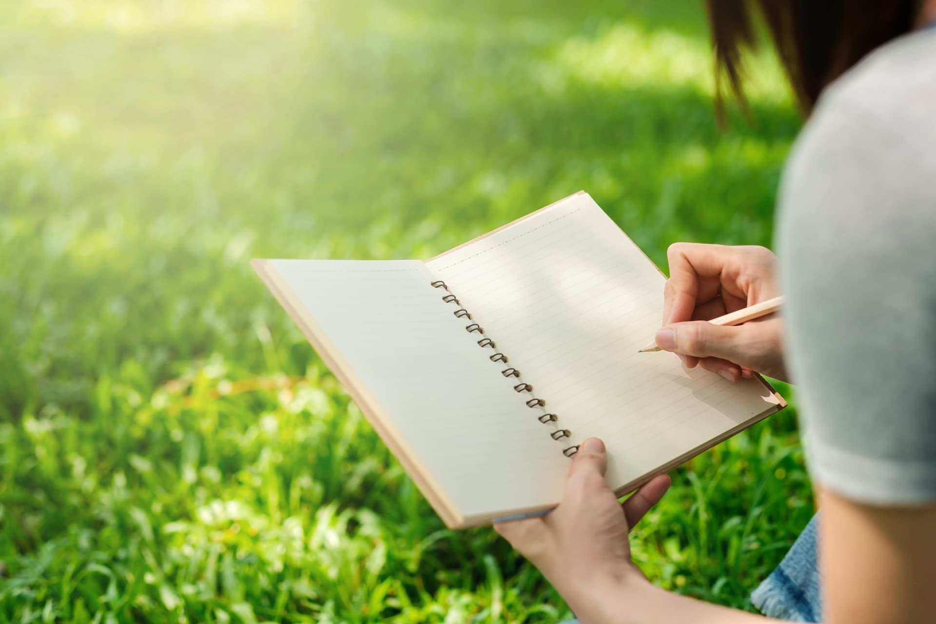 Person writing in notebook with pencil outdoors on green grass.