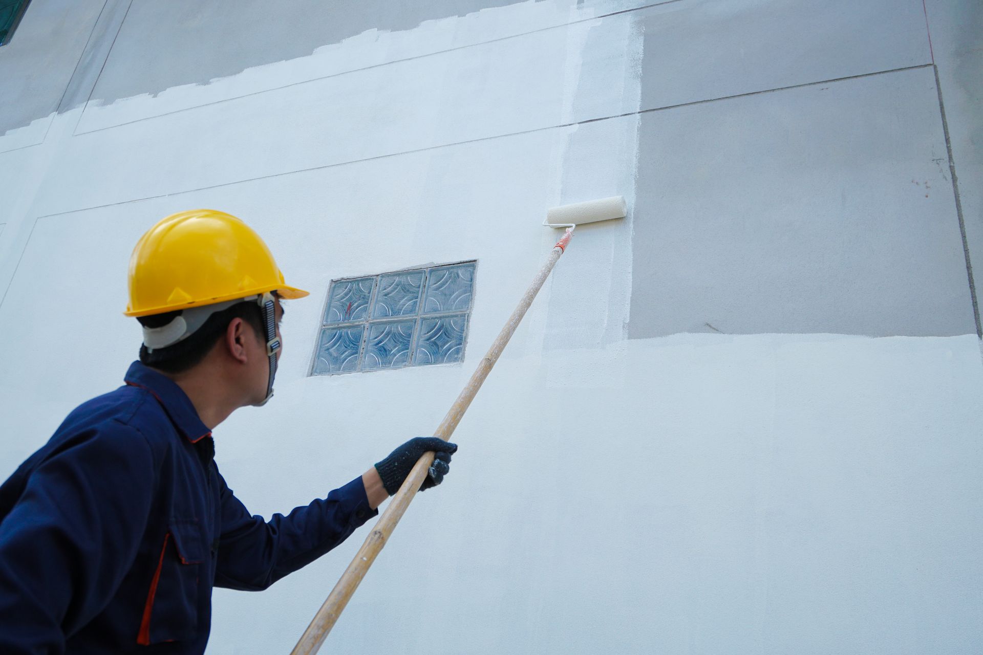 Person in a yellow hard hat painting an exterior wall with a roller.