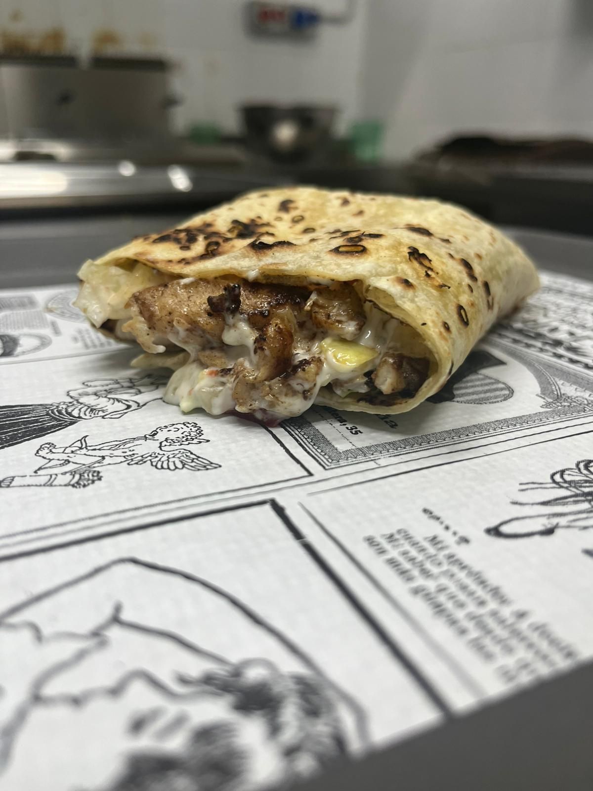 Mezzo burrito grigliato su un tavolo ricoperto di giornali, con il ripieno ben visibile; la cucina sullo sfondo è sfocata.