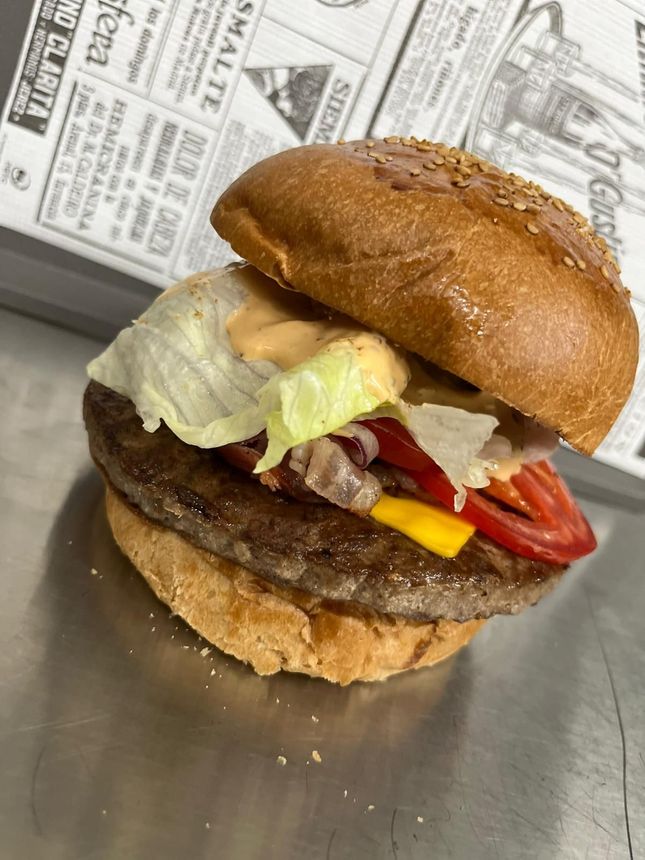 Primo piano di un cheeseburger con lattuga e pomodoro su una superficie metallica, con un giornale sullo sfondo.