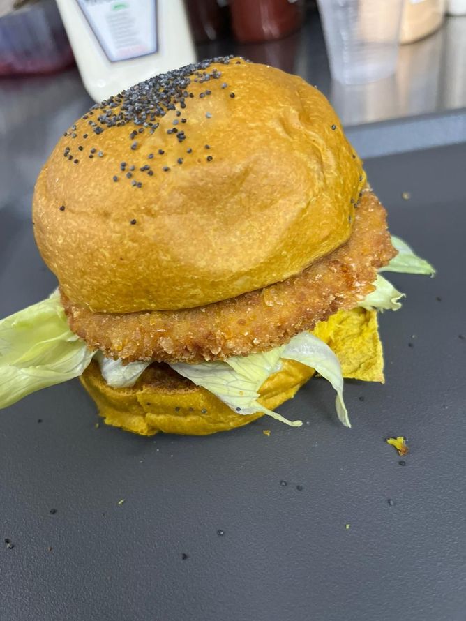 Hamburger con lattuga e salsa su un vassoio scuro, sormontato da un panino cosparso di semi di sesamo nero.