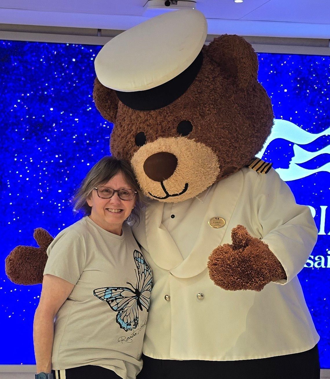 Woman smiles, poses with a large teddy bear in a sailor's uniform. Blue backdrop with white stars.