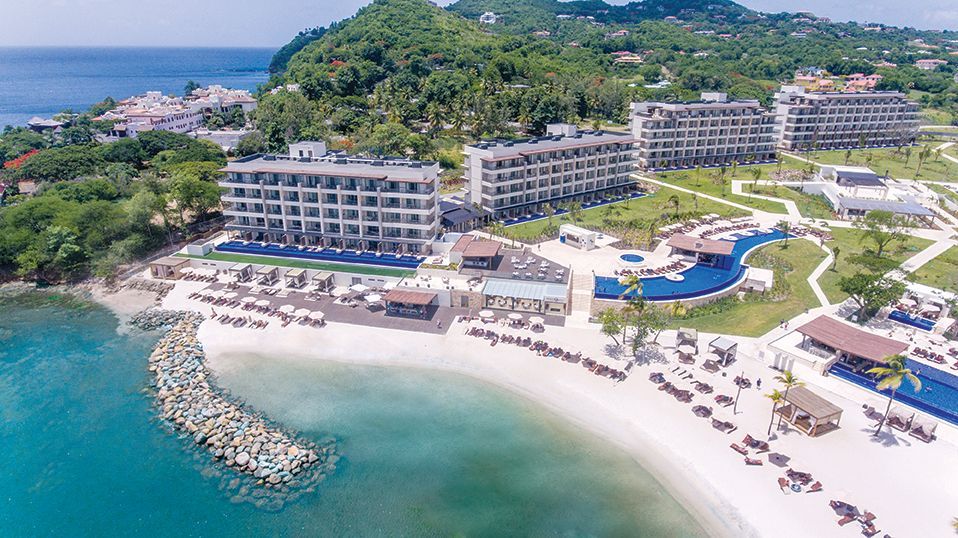 Beachfront resort with multiple gray buildings, pools, and sandy beach; green hills in the background.