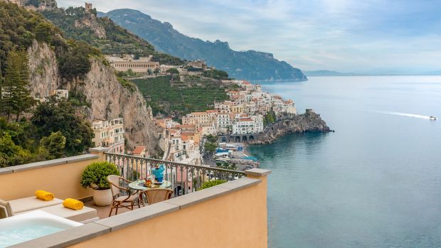 A sunny balcony with a hot tub overlooks the town of Amalfi, Italy, nestled on a cliffside by the Mediterranean Sea.