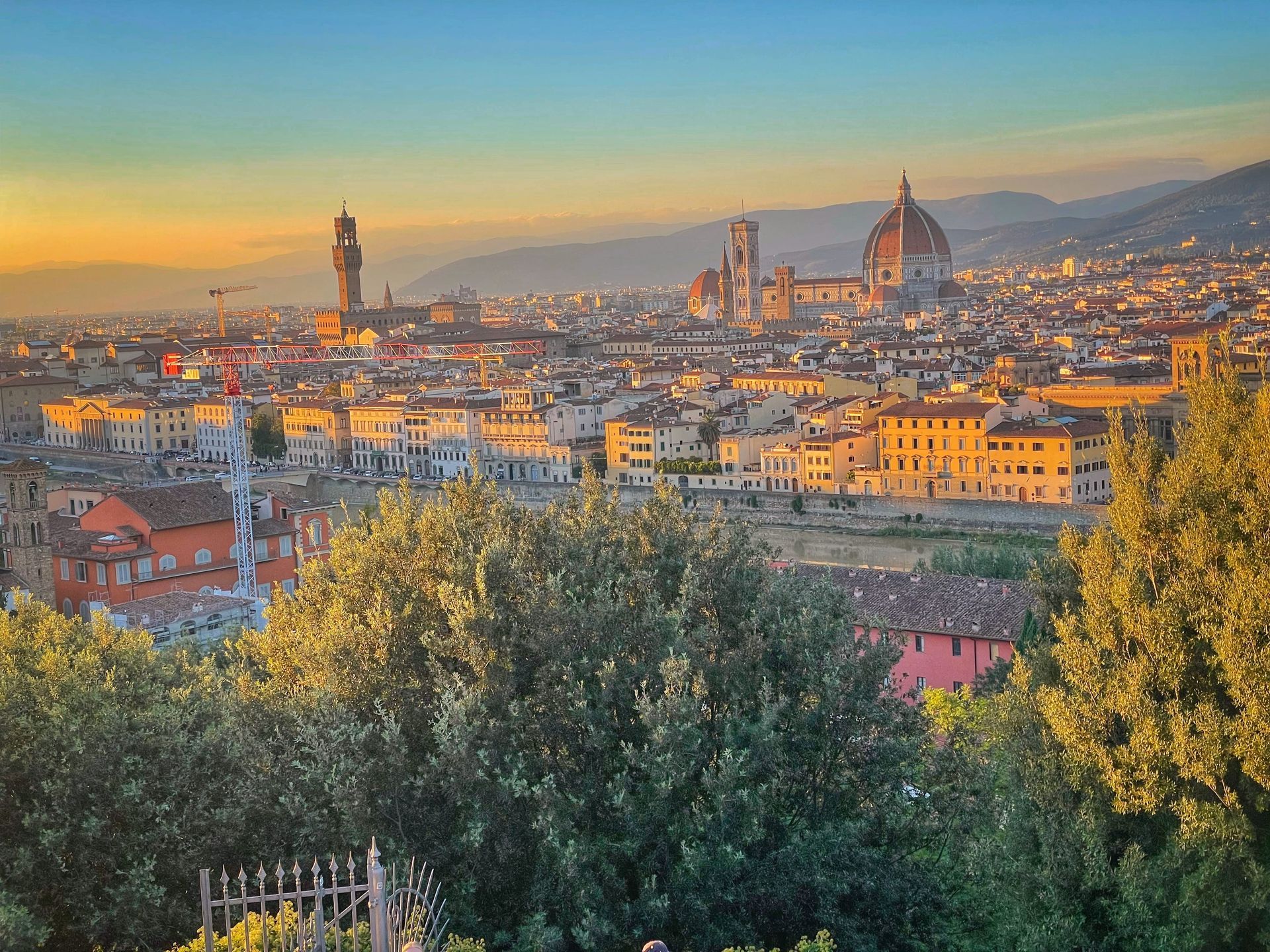 Panoramic sunset view over Florence, Italy, featuring the Duomo and Palazzo Vecchio rising above city buildings and trees.