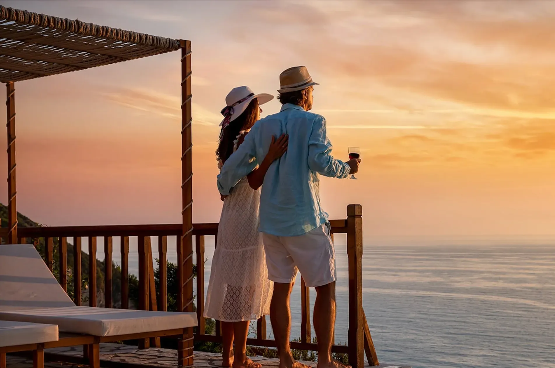 Couple on a wooden deck overlooking the ocean at sunset; the man holds a glass of wine.