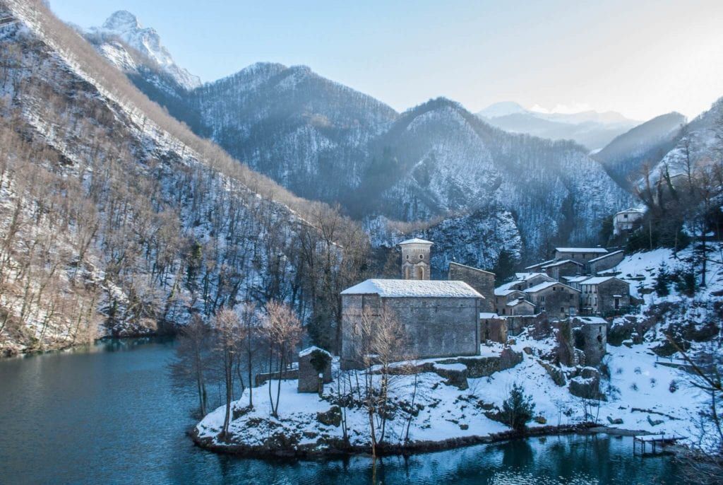 Snow-covered mountains surround a lake and stone buildings, including a church.