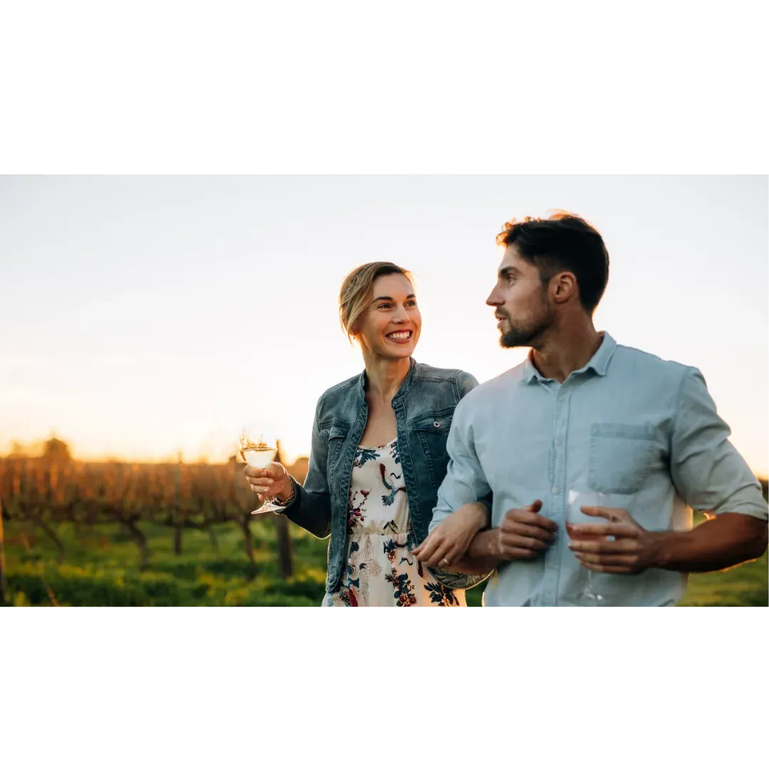 A couple holding wine glasses walk together through a vineyard at sunset.