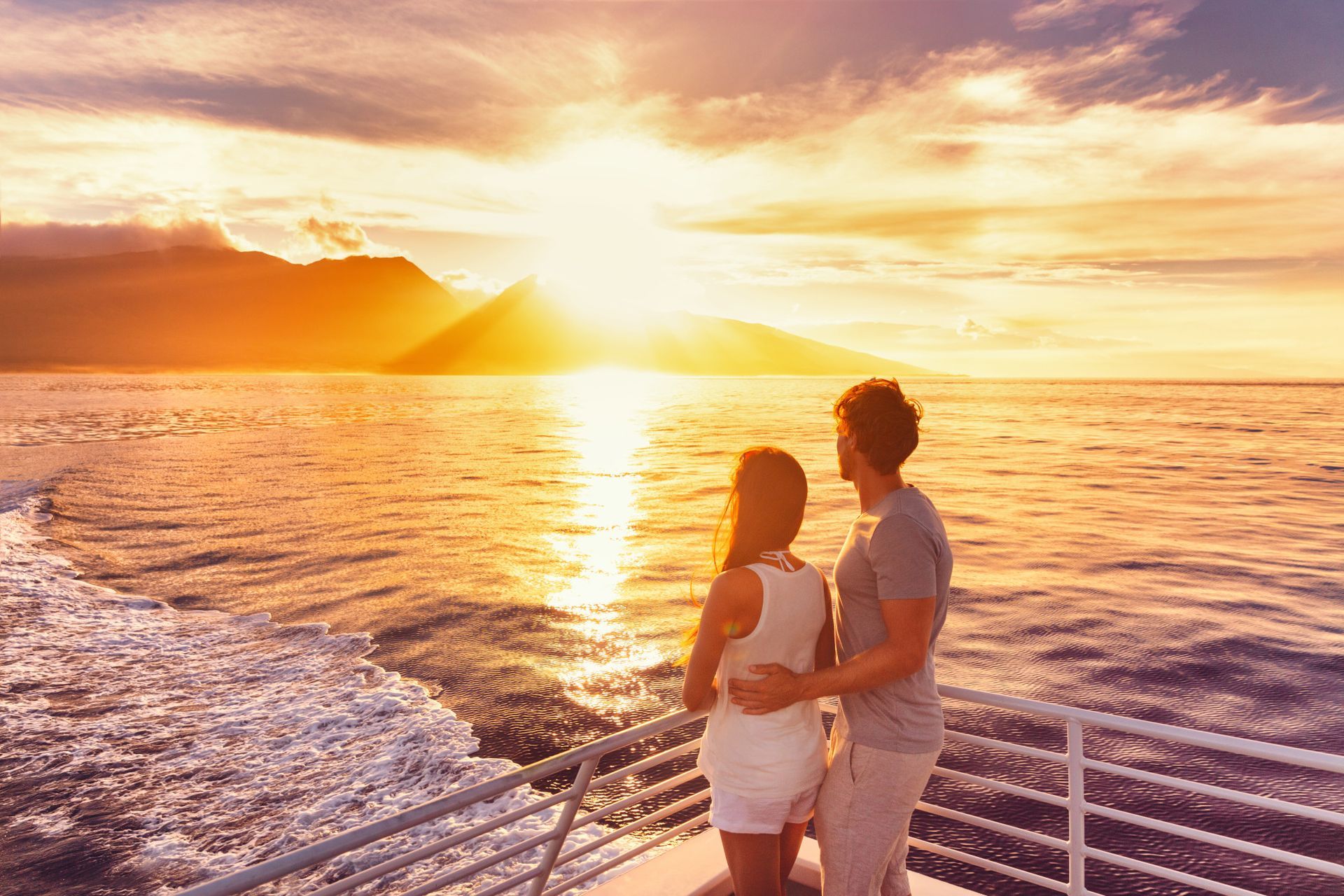 Couple on boat, watching sunset over ocean; sun reflecting on water, mountain in background.