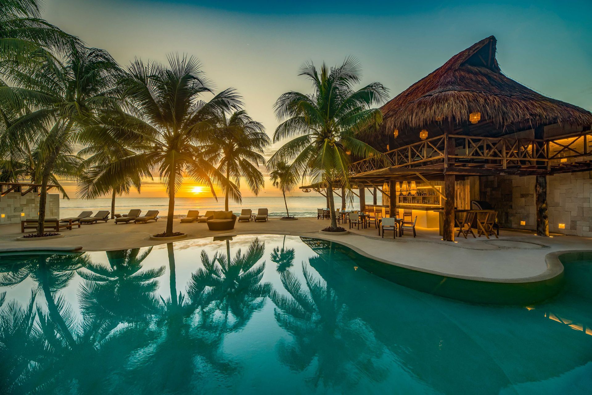 Tropical beach resort at sunset with pool, palm trees, and thatched-roof bar.
