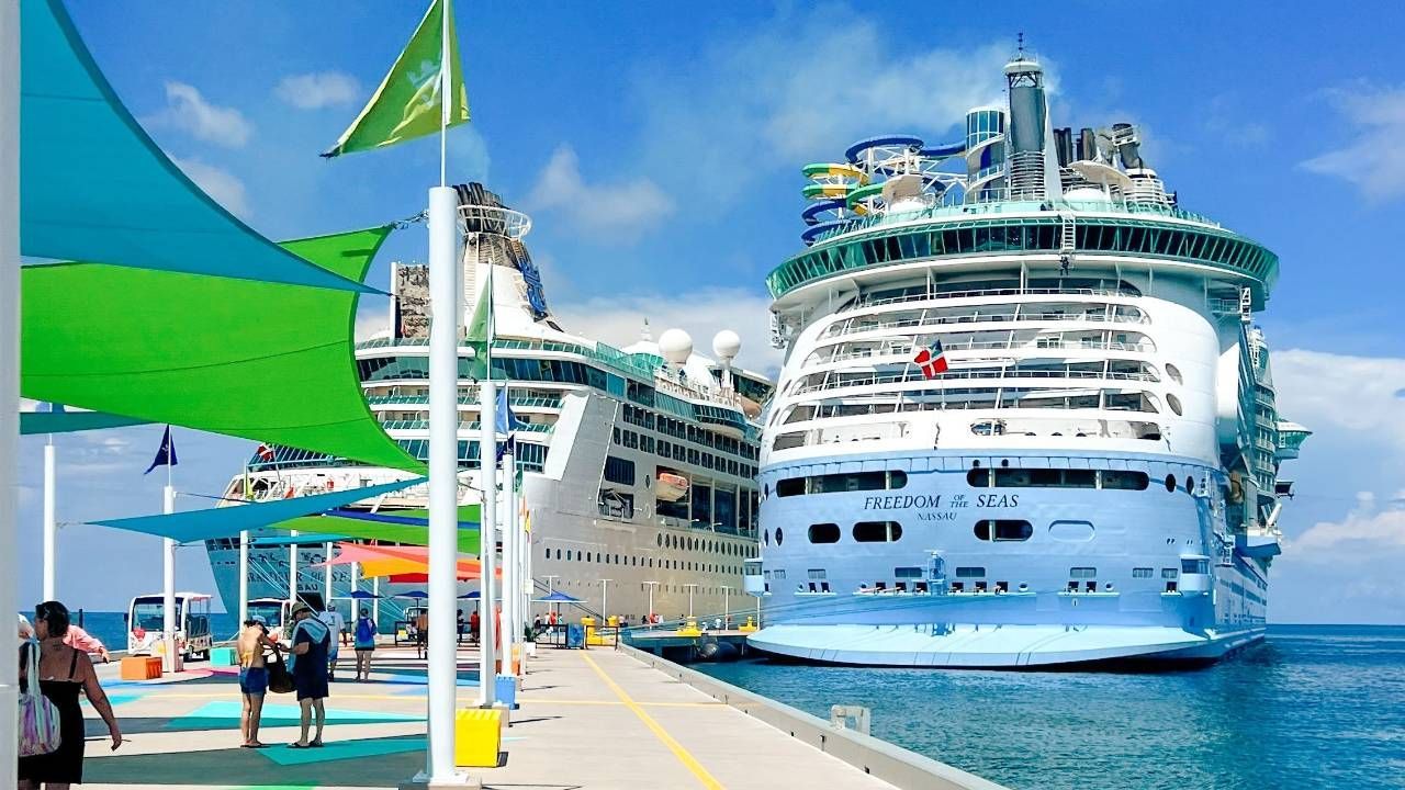 Two large cruise ships docked at a pier, people nearby; blue water and sky.