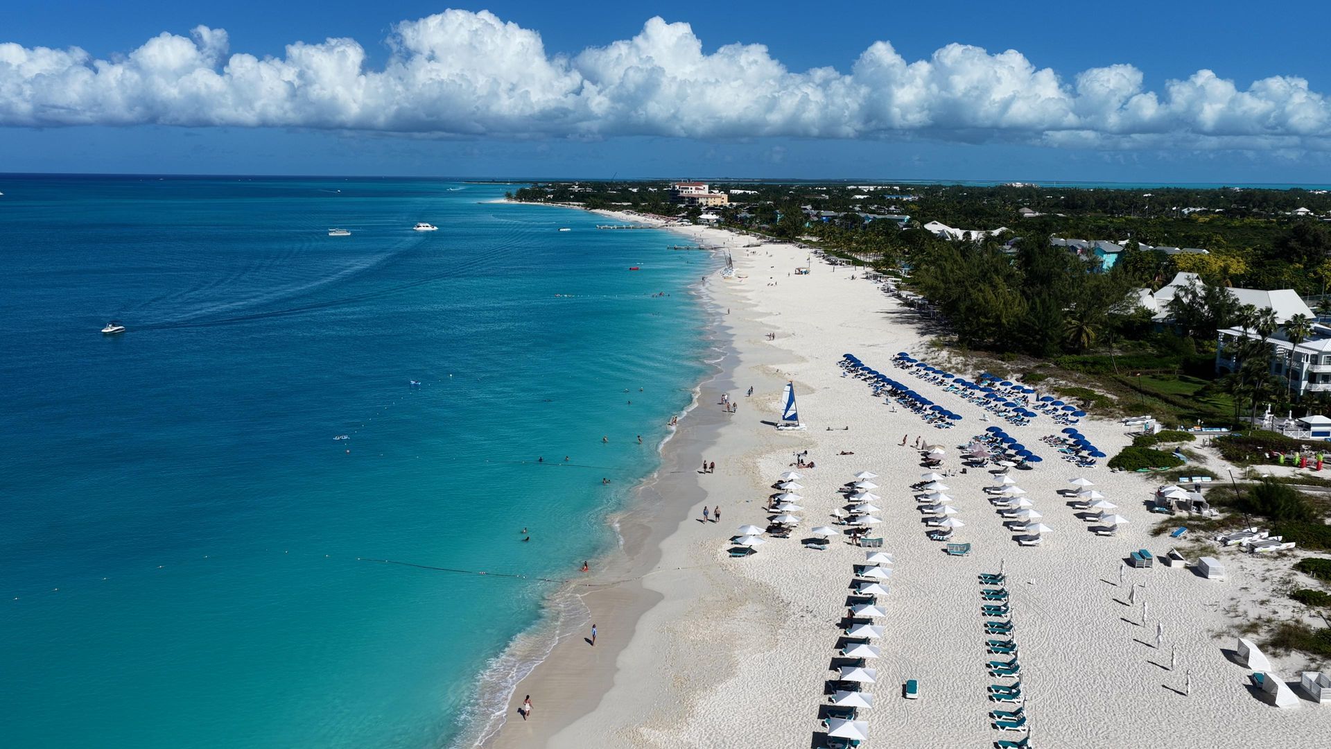 Aerial view of a white sand beach with turquoise water. Beach chairs, umbrellas, and buildings line the shore under a blue sky.