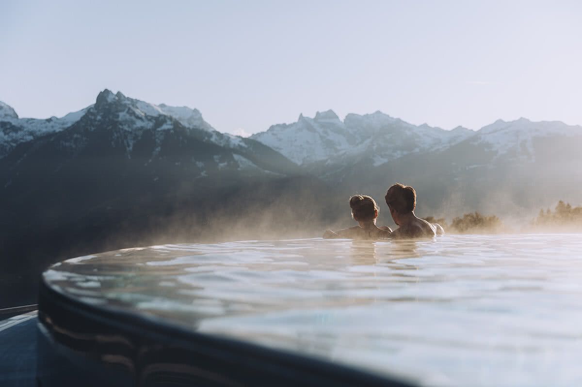 Two people sitting in an outdoor infinity pool looking out at snow-capped mountains at sunrise.