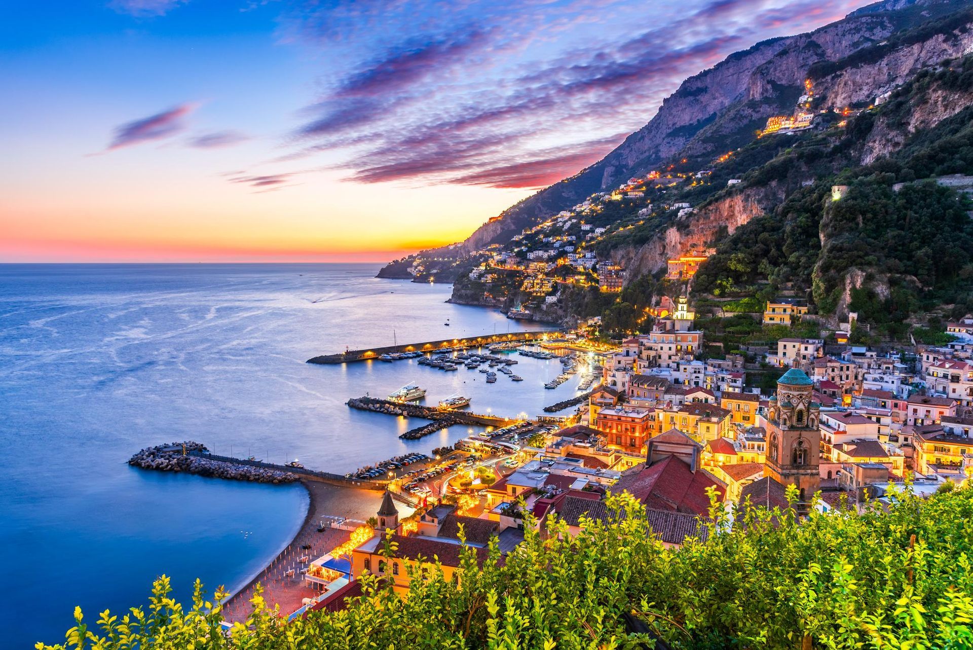 Coastal Italian town at sunset with buildings, boats, and a mountain along the water.