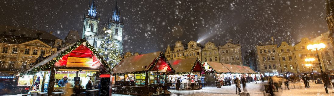 Christmas market at night, snow falling. Tower in background, illuminated stalls below, people.