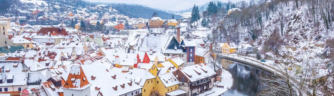 Snow-covered town with red-roofed buildings and a winding river, likely Cesky Krumlov, Czech Republic.
