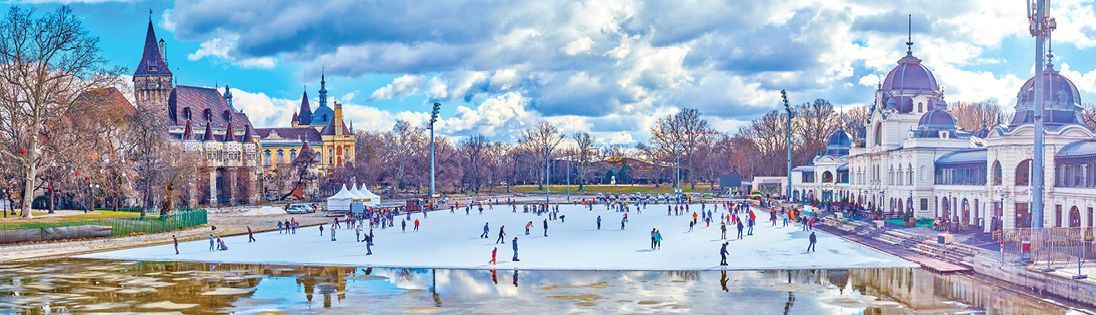 People ice skating on a frozen lake in a park, with buildings on either side. Cloudy blue sky.