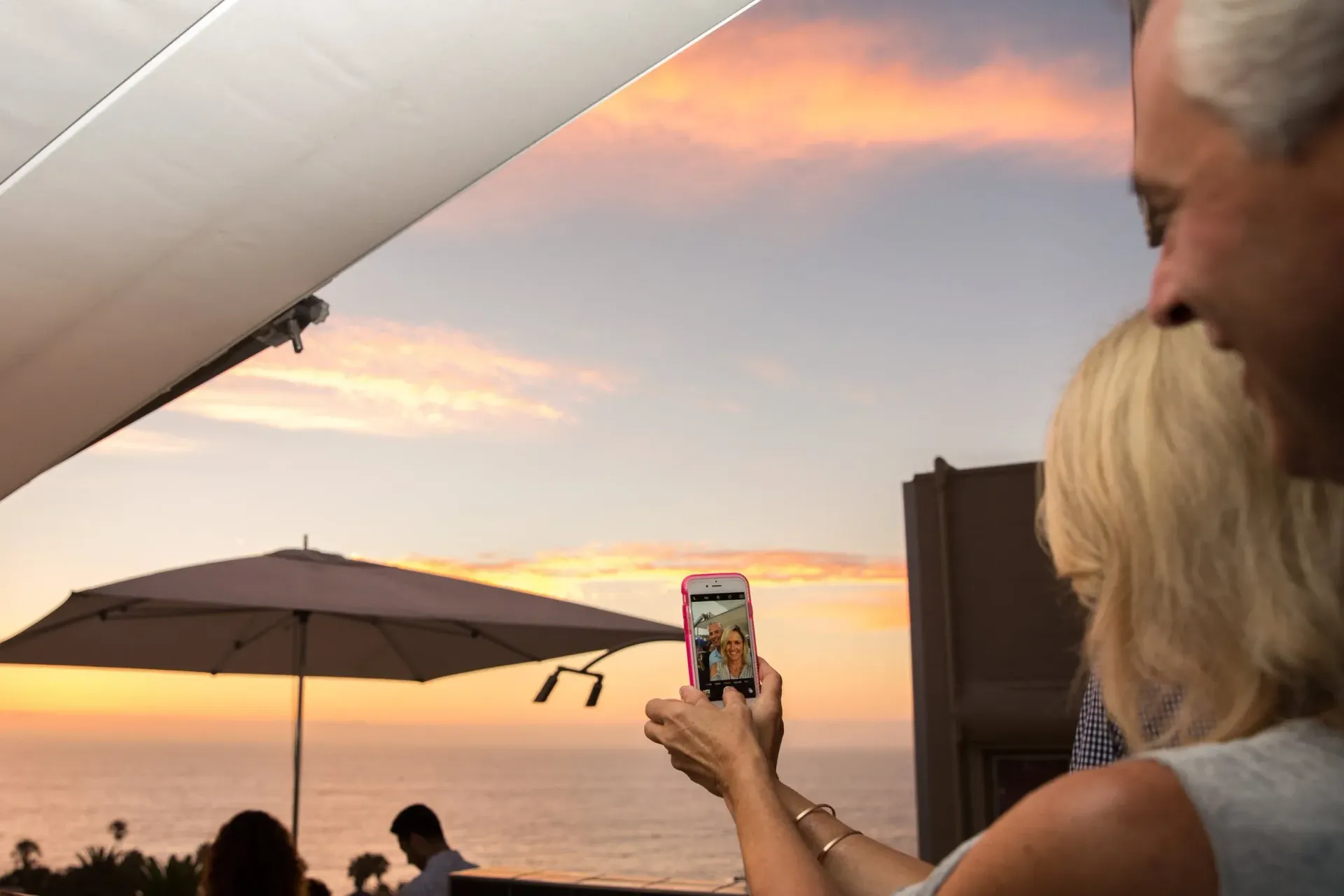 Woman taking a photo of a sunset over the ocean with a smartphone, viewed from a patio.