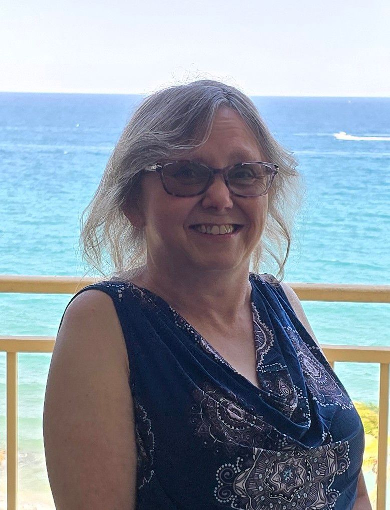 Woman wearing glasses smiles outdoors near ocean, blue dress, shoulder-up.