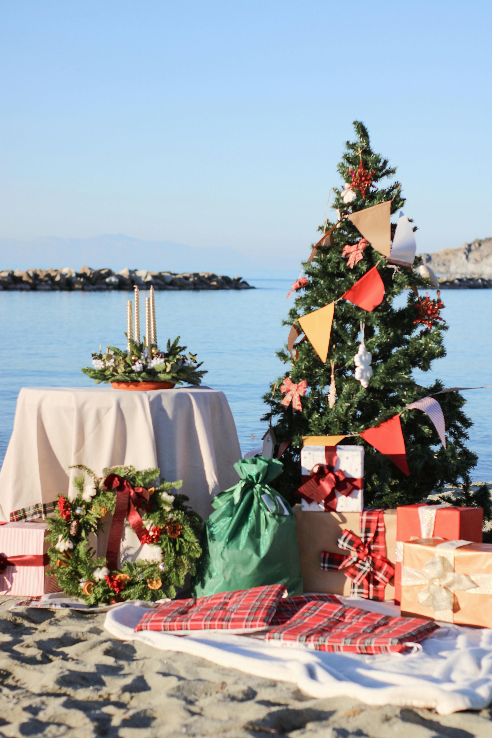 Christmas scene at the beach: tree and presents, table with candles, wreath. Blue sky and water.