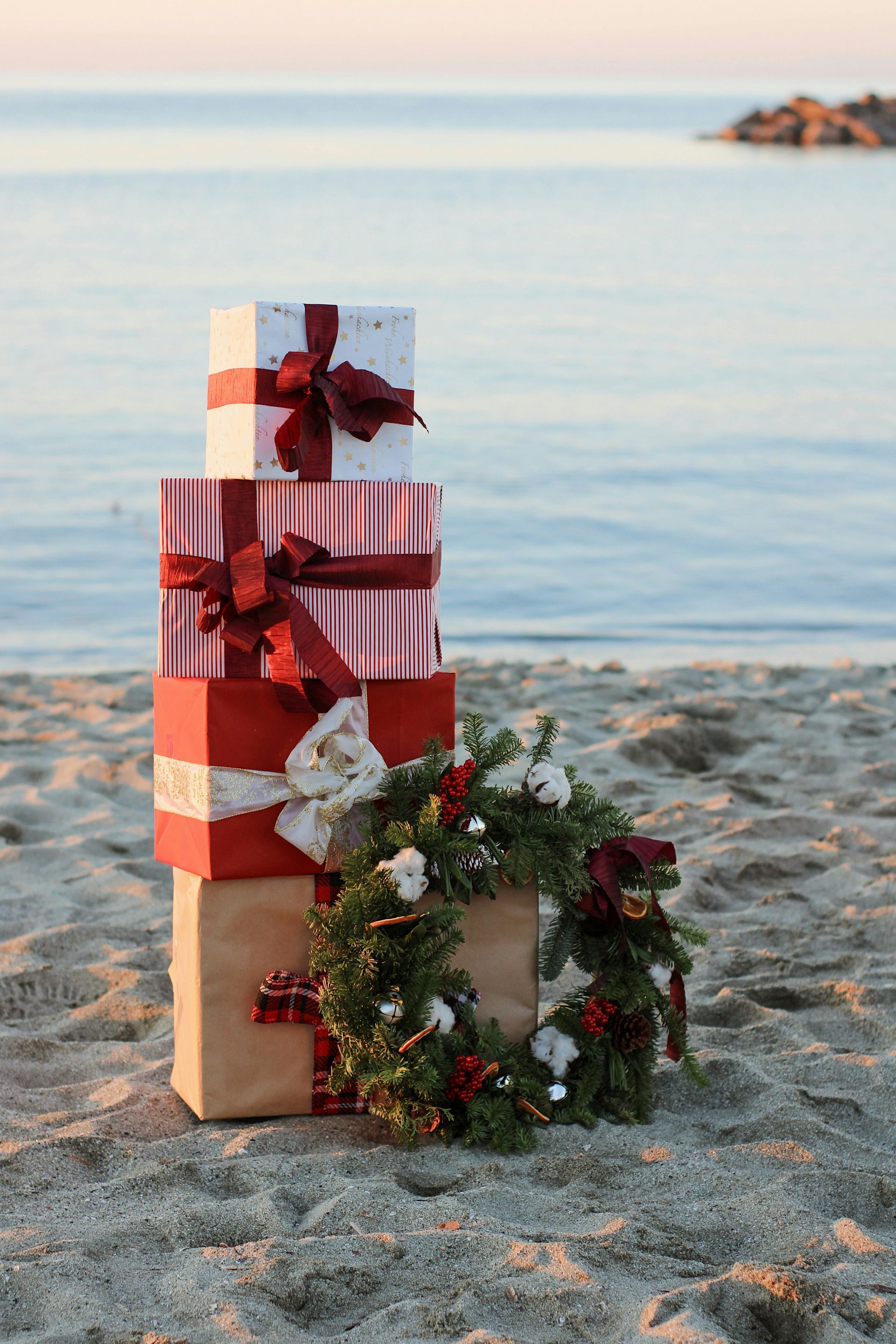 Stacked gifts on a beach, topped with a wreath, with the ocean in the background.