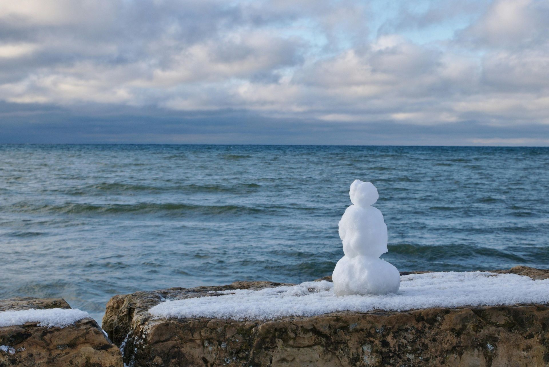 Snowman on a stone wall with a lake and cloudy sky in the background.